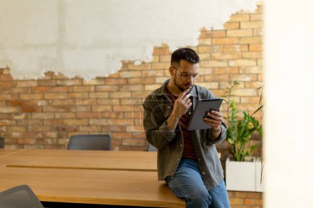Téléchargez les photos : Un homme joyeux tient avec confiance une tablette numérique dans un espace de bureau contemporain avec un mur de briques apparentes, symbolisant un mélange de technologie moderne et de charme rustique - en image libre de droit