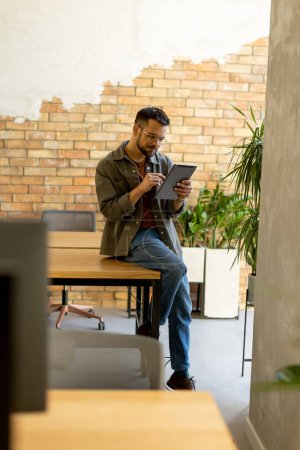 Hombre alegre se para con confianza sosteniendo una tableta digital en un espacio de oficina contemporáneo con una pared de ladrillo visto, que simboliza una mezcla de tecnología moderna y encanto rústico