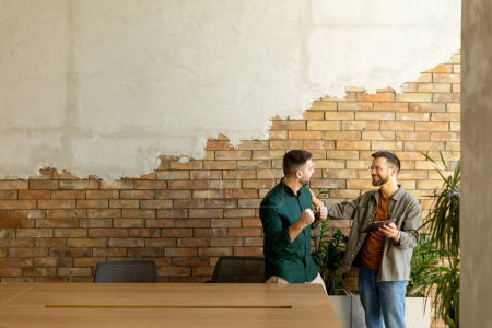 Photo for Two professionals are engaged in a focused conversation while holding digital tablet in a contemporary office space, featuring rustic exposed brickwork and warm natural light - Royalty Free Image