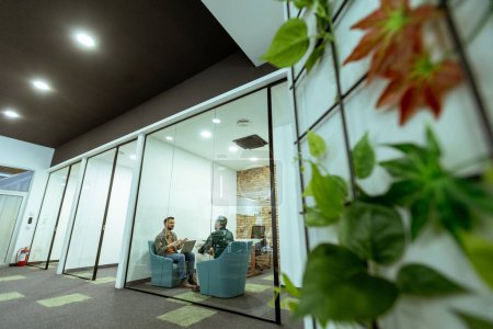 Two professionals engage in a conversation while seated comfortably in an office lounge, surrounded by greenery and contemporary design elements
