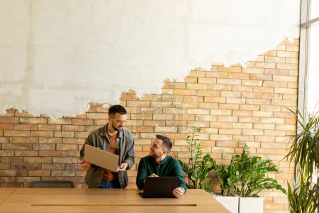 Photo for Two smiling professionals engage in a collaborative work session at a wooden table, their camaraderie evident in a contemporary office setting with an exposed brick wall backdrop - Royalty Free Image