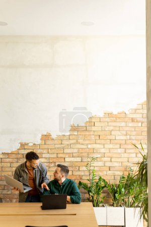 Two smiling professionals engage in a collaborative work session at a wooden table, their camaraderie evident in a contemporary office setting with an exposed brick wall backdrop