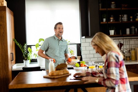 Téléchargez les photos : Femme lit depuis une tablette au comptoir de la cuisine tandis qu'un homme tenant une tomate lui sourit - en image libre de droit