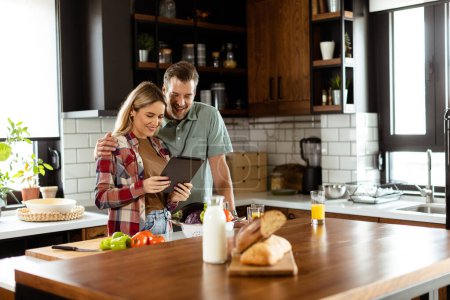 A cheerful couple stands in a well-lit kitchen, engrossed in a digital tablet among fresh ingredients