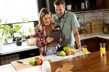 A cheerful couple stands in a well-lit kitchen, engrossed in a digital tablet among fresh ingredients