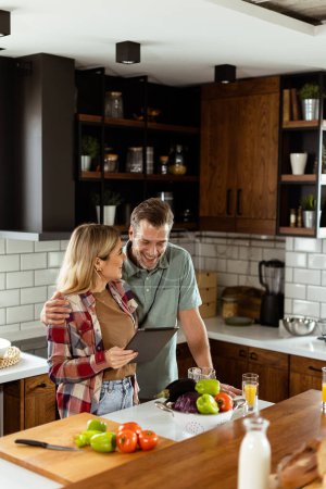 Foto de Una alegre pareja se encuentra en una cocina bien iluminada, absorta en una tableta digital entre ingredientes frescos - Imagen libre de derechos