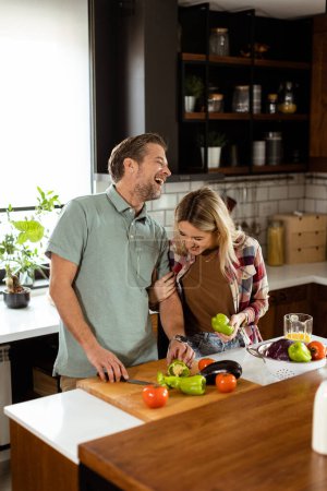 Foto de Un hombre y una mujer sonrientes cortando verduras frescas en una isla de cocina, disfrutando de una actividad de cocina saludable juntos - Imagen libre de derechos