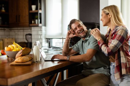Ein fröhliches Paar genießt einen unbeschwerten Moment in seiner sonnigen Küche und arbeitet am Laptop, umgeben von einem gesunden Frühstück