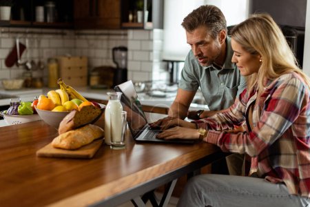 Foto de Una pareja alegre disfruta de un momento de luz en su cocina soleada, trabajando en un ordenador portátil rodeado de un desayuno saludable - Imagen libre de derechos
