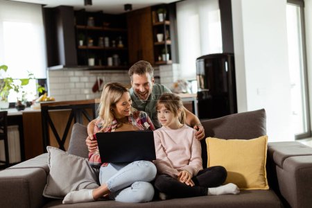 Téléchargez les photos : Joyeux famille de trois passe du temps ensemble sur le canapé du salon, partageant un moment autour d'un ordinateur portable dans leur maison confortable - en image libre de droit