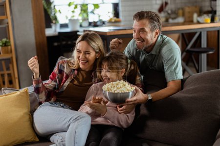 Photo for A family of three is comfortably nestled on a couch, their faces reflecting excitement and attentiveness as they share a bowl of popcorn during a suspenseful movie night - Royalty Free Image