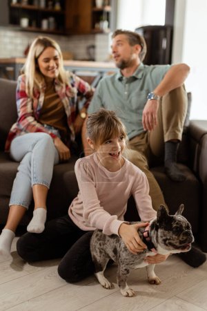 Téléchargez les photos : Des parents joyeux regardent leur fille jouer avec un bouledogue français heureux à la maison - en image libre de droit