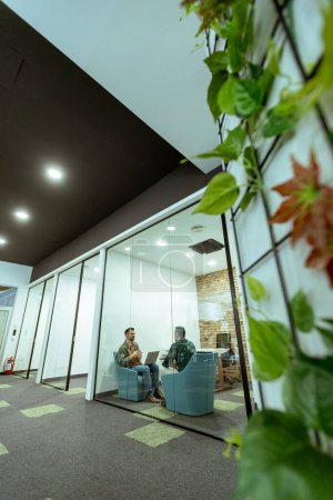 Deux professionnels s'engagent dans une conversation tout en s'asseyant confortablement dans un salon de bureau, entouré de verdure et d'éléments de design contemporain