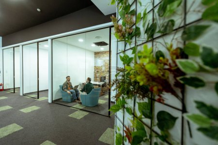 Dos profesionales conversan sentados cómodamente en un salón de oficina, rodeado de vegetación y elementos de diseño contemporáneo