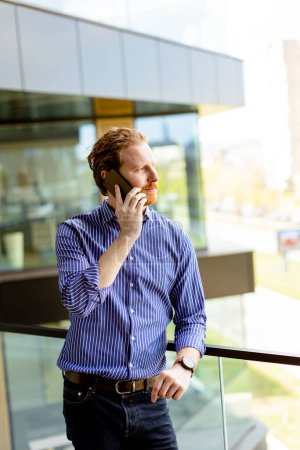 Foto de Un hombre enfocado en una camisa a rayas habla en su teléfono mientras domina el balcón a la luz del sol. - Imagen libre de derechos