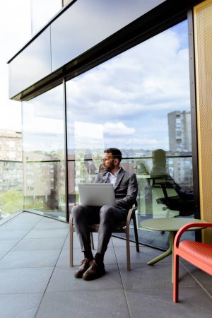 Téléchargez les photos : Un homme concentré en costume utilise son ordinateur portable sur un balcon ensoleillé surplombant le paysage urbain. - en image libre de droit