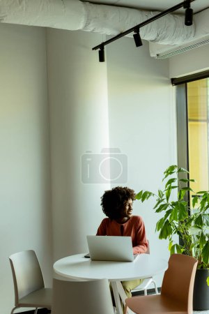 Foto de Una mujer alegre habla en su teléfono en una mesa de café con un ordenador portátil y una planta vibrante a su lado. - Imagen libre de derechos