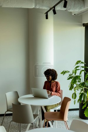 Photo for A cheerful woman talks on her phone at a cafe table with a laptop and a vibrant plant beside her. - Royalty Free Image