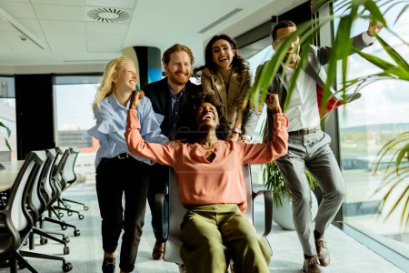 Foto de Equipo de compañeros de trabajo comparte un momento de diversión, corriendo una silla de oficina en medio de un espacio de trabajo bien iluminado. - Imagen libre de derechos