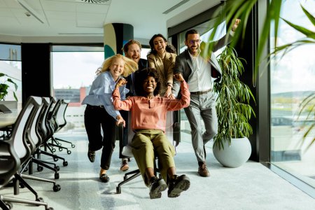 Foto de Equipo de compañeros de trabajo comparte un momento de diversión, corriendo una silla de oficina en medio de un espacio de trabajo bien iluminado. - Imagen libre de derechos