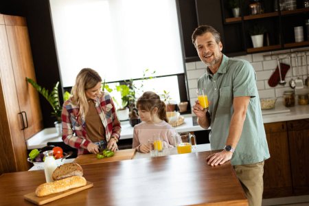Téléchargez les photos : Un homme joyeux avec un verre de jus d'orange tandis qu'une femme et un enfant préparent la nourriture dans une cuisine confortable. - en image libre de droit