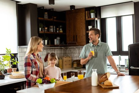 Téléchargez les photos : Une famille joyeuse profite d'une conversation chaleureuse dans leur cuisine avec du jus de fruits frais et un ordinateur portable assis sur la table. - en image libre de droit