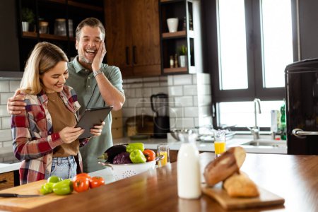 Ein fröhliches Paar steht in einer gut beleuchteten Küche, vertieft in ein digitales Tablet zwischen frischen Zutaten