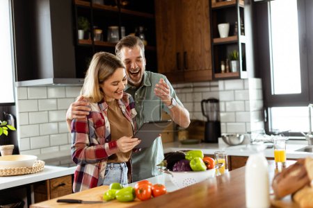 Foto de Una alegre pareja se encuentra en una cocina bien iluminada, absorta en una tableta digital entre ingredientes frescos - Imagen libre de derechos