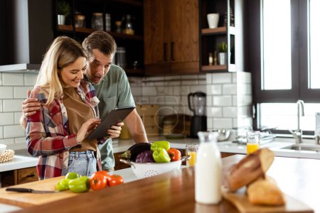 Ein fröhliches Paar steht in einer gut beleuchteten Küche, vertieft in ein digitales Tablet zwischen frischen Zutaten