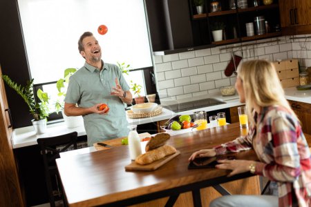 Foto de La mujer lee de una tableta en el mostrador de la cocina mientras un hombre sosteniendo un tomate le sonríe - Imagen libre de derechos
