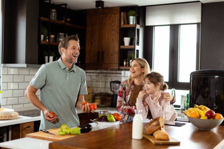 Téléchargez les photos : Jeune famille bavardant et préparant la nourriture autour d'un comptoir de cuisine animé rempli d'ingrédients frais et d'ustensiles de cuisine - en image libre de droit