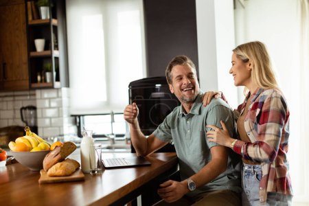 Photo for A cheerful couple enjoys a light-hearted moment in their sunny kitchen, working on laptop surrounded by a healthy breakfast - Royalty Free Image