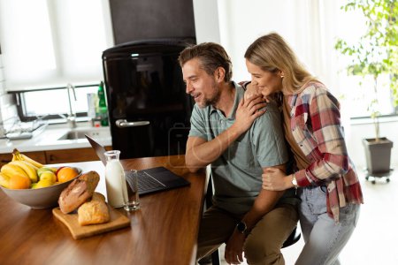 A cheerful couple enjoys a light-hearted moment in their sunny kitchen, working on laptop surrounded by a healthy breakfast