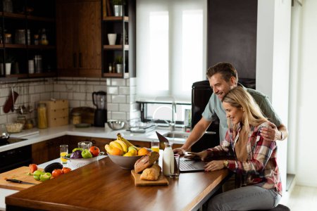 Ein fröhliches Paar genießt einen unbeschwerten Moment in seiner sonnigen Küche und arbeitet am Laptop, umgeben von einem gesunden Frühstück