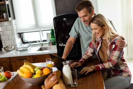 Foto de Una pareja alegre disfruta de un momento de luz en su cocina soleada, trabajando en un ordenador portátil rodeado de un desayuno saludable - Imagen libre de derechos