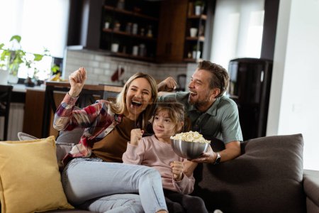 Photo for A family of three is comfortably nestled on a couch, their faces reflecting excitement and attentiveness as they share a bowl of popcorn during a suspenseful movie night - Royalty Free Image
