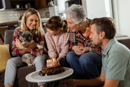 Téléchargez les photos : Scène chaleureuse se déroule comme une famille multi-générationnelle se réunit sur un canapé pour présenter un gâteau d'anniversaire à une grand-mère ravie, créant des souvenirs à chérir - en image libre de droit
