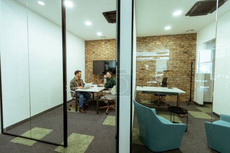 Foto de Dos profesionales conversan sentados cómodamente en un salón de oficina, rodeado de vegetación y elementos de diseño contemporáneo - Imagen libre de derechos
