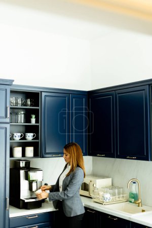 Une femme souriante en tenue de travail tenant une tasse à café, prenant une pause détente dans une cuisine bleue élégante.