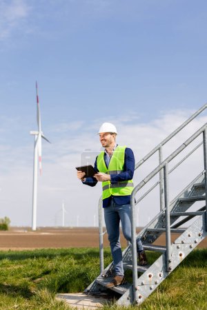 Foto de Un ingeniero alegre en equipo de seguridad revisa los datos en una tableta antes de elevar los molinos de viento bajo un cielo despejado. - Imagen libre de derechos