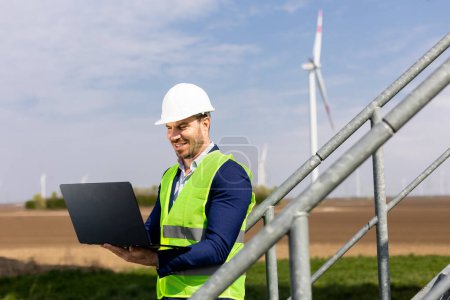 Photo for A focused engineer examines his laptop with a wind turbine in the background under a clear sky. - Royalty Free Image