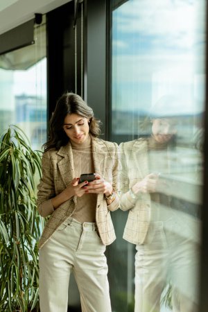 Téléchargez les photos : Femme élégante regarde par la fenêtre, tenant un smartphone, au milieu de l'allure urbaine - en image libre de droit