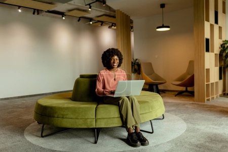Téléchargez les photos : Femme d'affaires afro-américaine spirituelle souriant tout en travaillant sur son ordinateur portable, confortablement assis dans un salon de bureau dynamique. - en image libre de droit