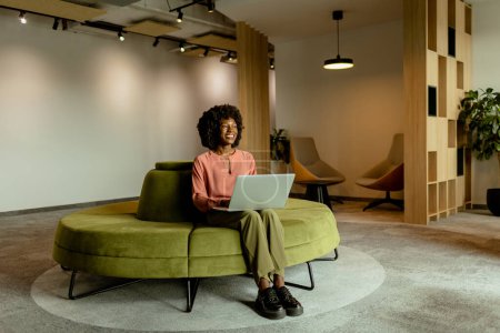 Téléchargez les photos : Femme d'affaires afro-américaine spirituelle souriant tout en travaillant sur son ordinateur portable, confortablement assis dans un salon de bureau dynamique. - en image libre de droit