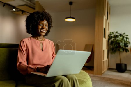 Foto de Mujer de negocios afroamericana animada sonriendo mientras trabaja en su computadora portátil, cómodamente sentada en un vibrante salón de oficina. - Imagen libre de derechos