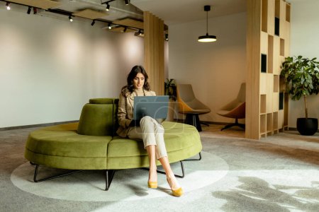Foto de La mujer enfocada trabaja en su computadora portátil, sentada en un elegante sofá verde en una oficina contemporánea bien iluminada - Imagen libre de derechos