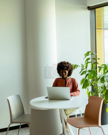 Téléchargez les photos : Une femme concentrée sur son ordinateur portable à une table de café lumineuse près d'une grande fenêtre à côté de plantes d'intérieur. - en image libre de droit