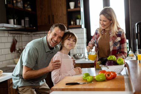 Téléchargez les photos : Jeune famille bavardant et préparant la nourriture autour d'un comptoir de cuisine animé rempli d'ingrédients frais et d'ustensiles de cuisine - en image libre de droit
