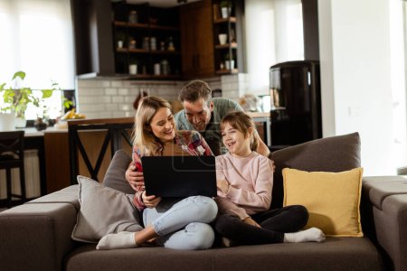 Photo for Joyful family of three spends quality time together on the living room sofa, sharing a moment around a laptop in their comfortable home - Royalty Free Image