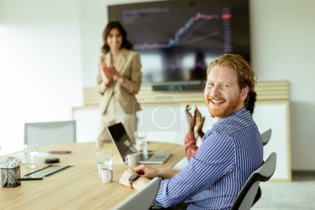 Foto de Hombre barbudo sonríe a la cámara mientras una mujer presenta un gráfico en una pantalla en una sala de reuniones moderna. - Imagen libre de derechos